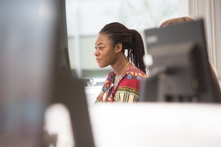 A black woman in a colourful shirt sat looking at a monitor