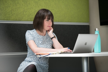 A white woman sat at a desk with her legs crossed looking at ipad screen and typing